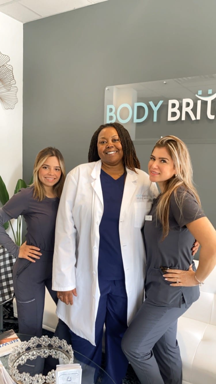 Three medical professionals—one in a white coat and two in grey scrub uniforms—stand smiling in front of a sign that reads