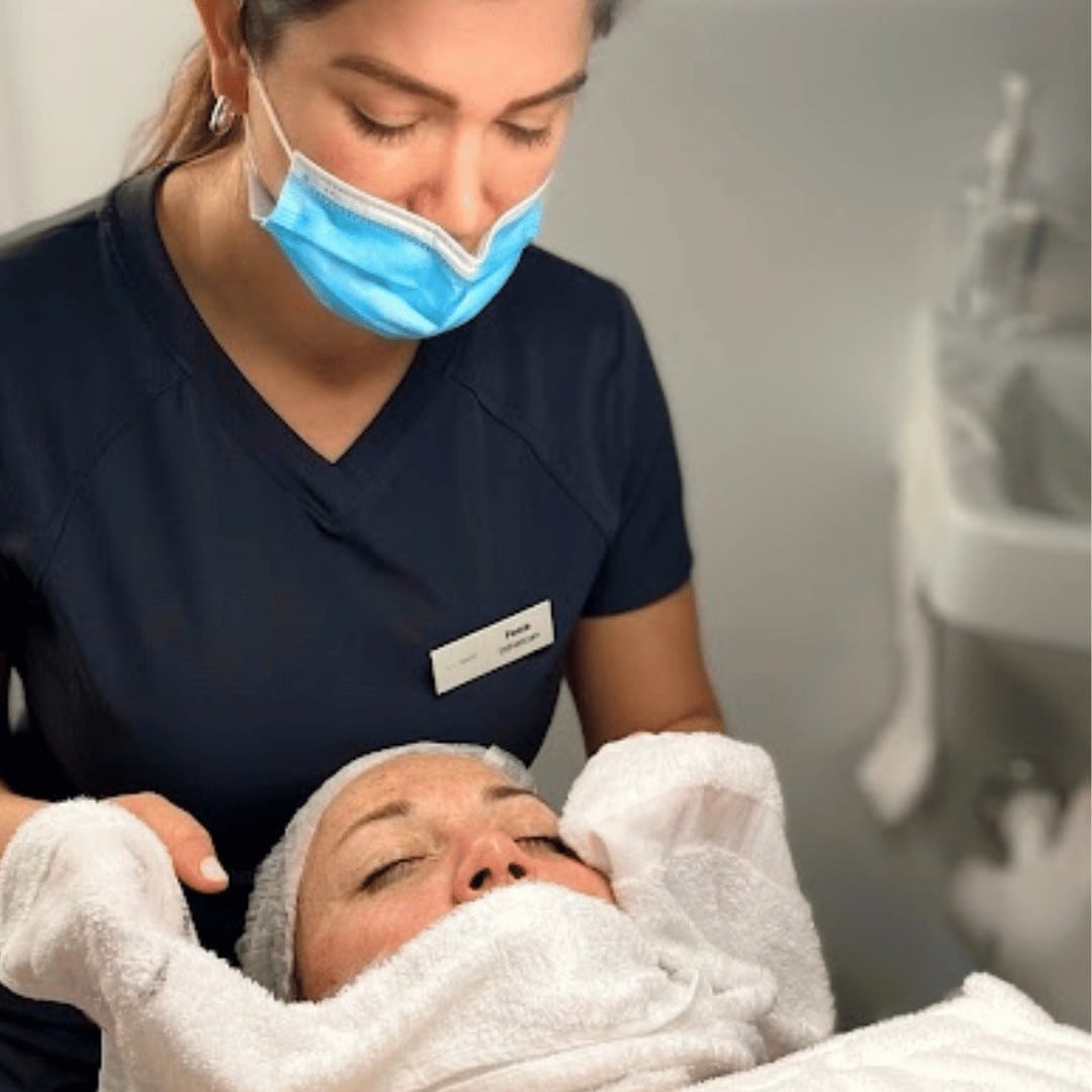 In a serene med spa located in Cooper City, a person clad in a blue uniform and mask meticulously performs a facial treatment on an individual who is lying down with their eyes closed, covered by a pristine white towel. This description encapsulates the tranquil ambiance of the setting perfectly.