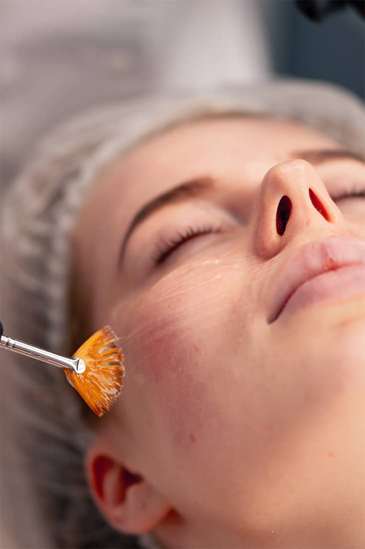 A person is enjoying a soothing facial treatment at a premier med spa in Cooper City. A brush gently applies a clear, rejuvenating substance to their cheek as they wear a disposable head cover and relax with their eyes closed, immersed in the tranquil ambiance of the top-rated local facility.