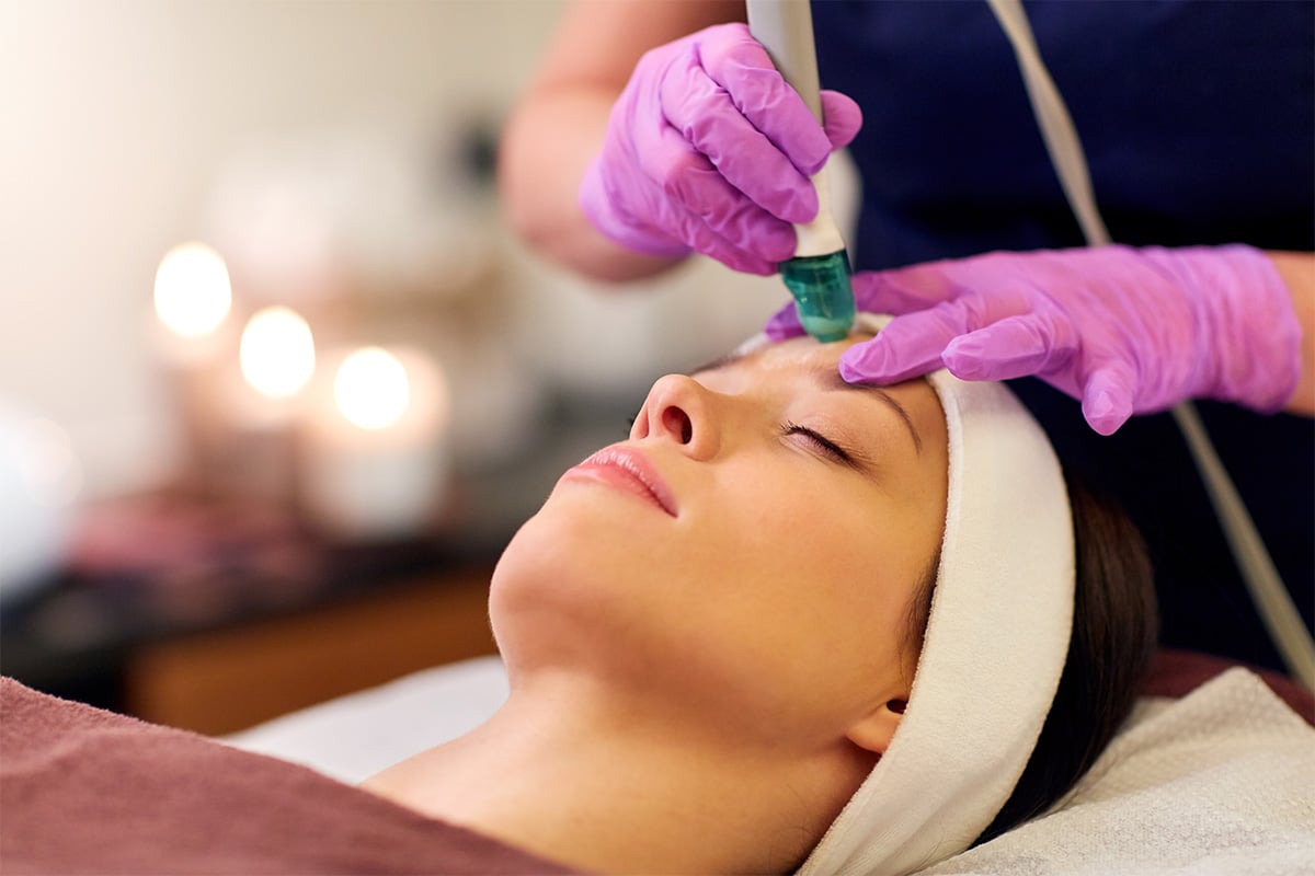 In a serene med spa setting in Cooper City, where lit candles provide a tranquil ambiance, a person lies down with their eyes closed, receiving an indulgent facial treatment. A professional, wearing purple gloves, meticulously applies a specialized tool on the individual's forehead to enhance their relaxation and skincare experience.