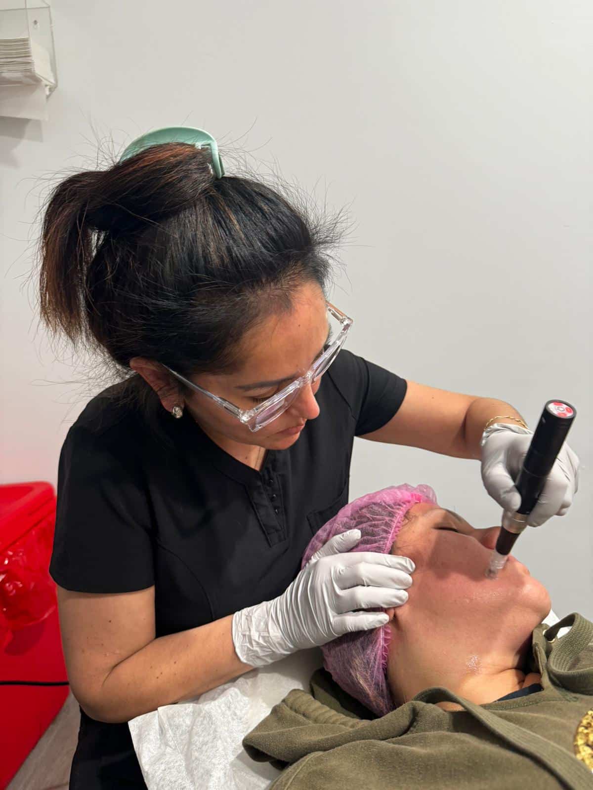 In a med spa located in Cooper City, a skilled female professional equipped with gloves and protective eyewear meticulously performs a microneedling procedure on a patient's face. The patient, whose hair is covered with a pink cap, benefits from the clinic's specialized skincare services designed to enhance and rejuvenate the skin.