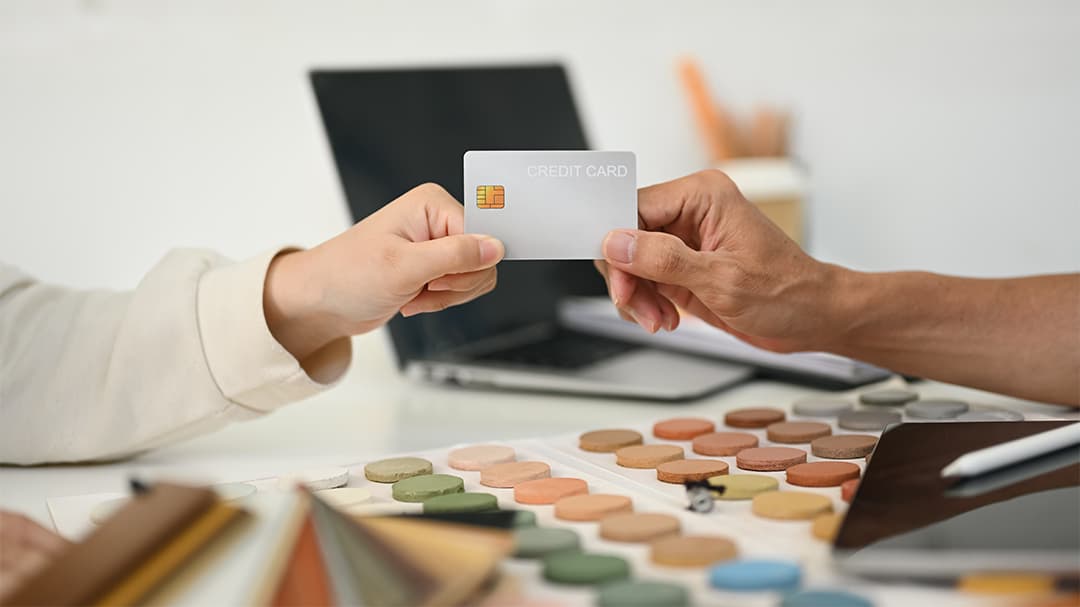 In a med spa in Cooper City, two people are exchanging a CareCredit card over a table adorned with an array of vibrant color swatches and a sleek laptop in the background.