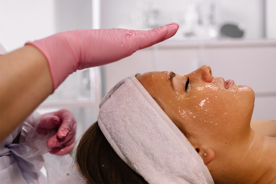 In a med spa located in Cooper City, a person with a facial mask lies down while another wearing pink gloves prepares to administer the Mesoestetic Cosmelan Peel, specifically targeting pigmentation and discoloration. The person receiving the treatment relaxes with closed eyes and a white headband securely in place.