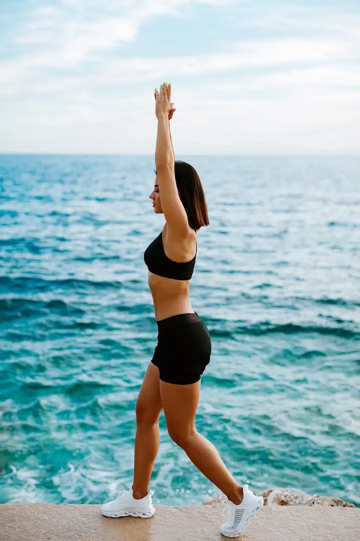 Dressed in athletic wear, the person poses near the ocean in Cooper City, arms raised triumphantly as they step forward, embodying strength and balance against the backdrop of blue water and clear sky. This empowering moment is akin to embarking on a transformative journey like Hormone Replacement Therapy (HRT), similar to rejuvenating experiences offered at local med spas. In Cooper City, these med spas provide innovative treatments that support personal transformation and well-being.