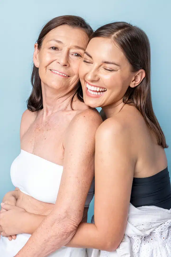 Against a blue background, two women share a warm embrace, exuding joy and vitality. Their smiles reflect the rejuvenating benefits of peptide therapy often found at med spas in Cooper City. The older woman, with vibrant shoulder-length brown hair, appears revitalized while the younger woman showcases her health with flowing long brown hair. Both wear sleeveless tops, embodying the essence of health and happiness that can be experienced through advanced wellness treatments available in Cooper City's renowned med spas.