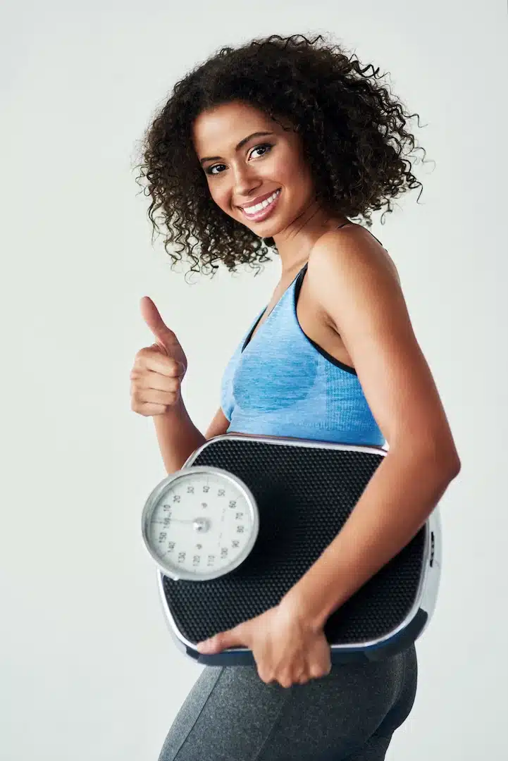 A woman in athletic wear smiles and gives a thumbs up, feeling confident in her progress with peptide therapy at a renowned med spa in Cooper City, while holding a bathroom scale.