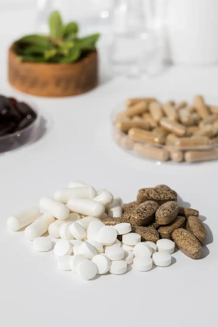 Amidst a pristine white surface, a collection of supplements, including neatly arranged white capsules and brown tablets, captures the essence of nutraceuticals. This scene echoes the modern aesthetic found in med spas across Cooper City, where wellness is enhanced through vitamin shots and advanced health therapies. A small plant and other indistinct items linger in the background, creating an atmosphere reminiscent of these innovative clinics dedicated to promoting holistic well-being.