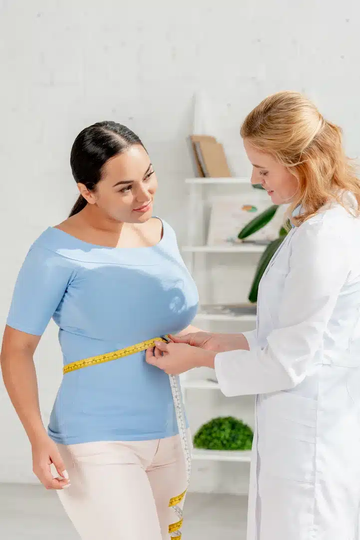 In a bright room at a med spa in Cooper City, a doctor carefully measures a woman's waist with a measuring tape, guiding her on her weight loss journey. She is dressed in a stylish blue shirt and white pants, embodying both fashion and determination as she commits to achieving her health goals.