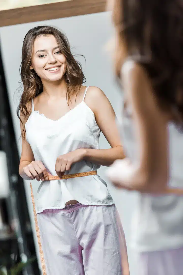 A woman in a white tank top and purple pants smiles with pride while marveling at her weight loss journey, measuring her waist with a tape in front of the mirror. Her success is partly thanks to the innovative treatments offered by a renowned med spa in Cooper City, where personalized wellness plans helped her achieve her goals.